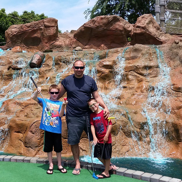 a dad and his two kids playing mini-gold with a waterfall behind them with blue water