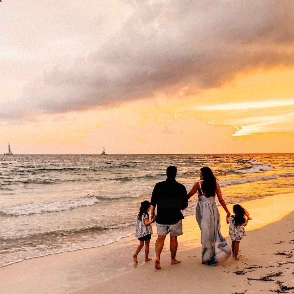 a mom dad and their two daughters all holding hands walking down the the shoreline of the beach during the sunset