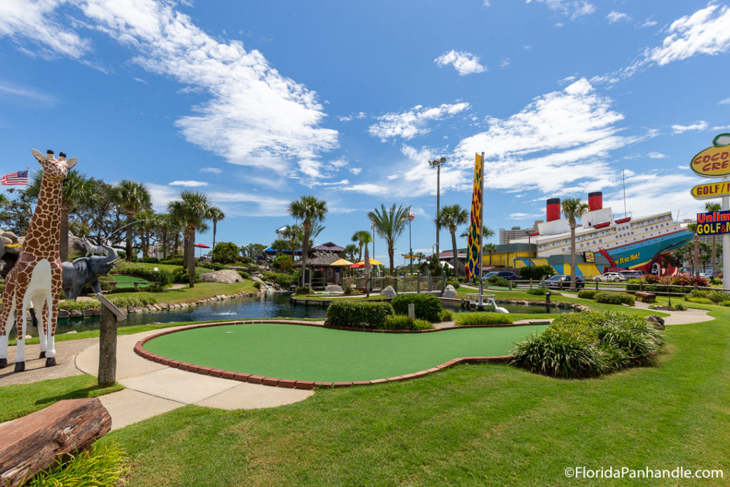 adventure mini-golf park with a giant boat in the back and a statue of a giraffe by the water next to the put hole