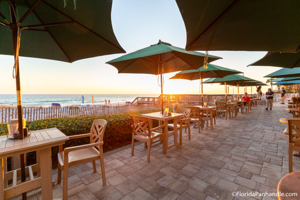 an outdoor dining area with tan colored seating and green umbrellas  during the sunset and a view of the beach