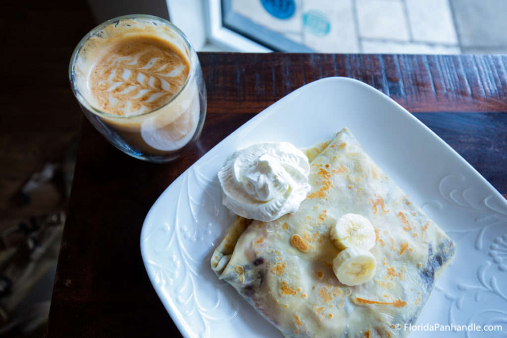 a dessert crepe with two banana slices on top and a side of whipped cream and a cup of coffee on the side with milk foam