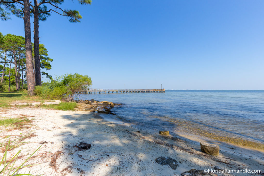 grassy beach area with trees and pier in the distance in the water at Mattie Kelly Park