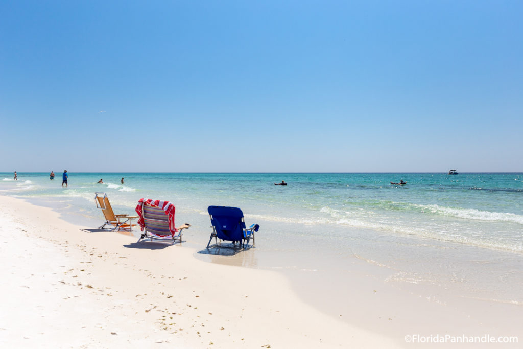 3 empty beach chairs along the shoreline and some people swimming in the water at Miramar Beach in Florida, best beaches florida panhandle