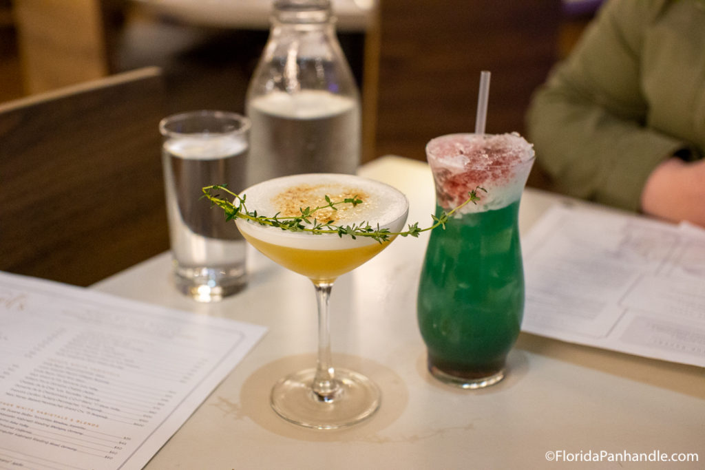 yellowish eggwhite drink with rosemary garnish on top inside coupe glass next to greenish drink with red sprinkles at the top, Upscale restaurants in destin