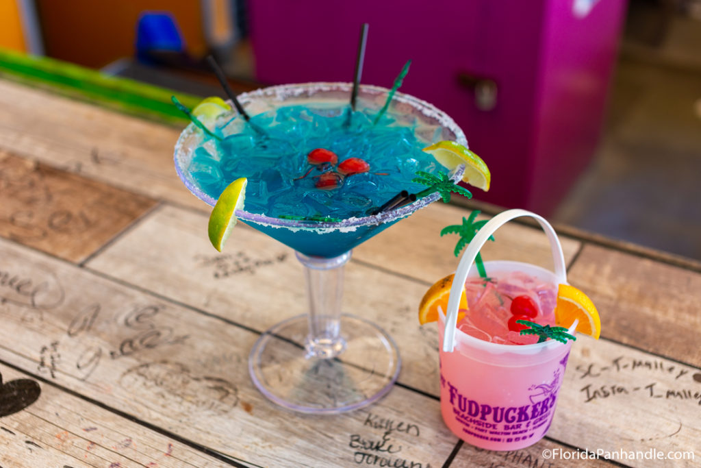 a tall glass with a blue drink in it and a small bucket filled with a pink drink with fruit in it