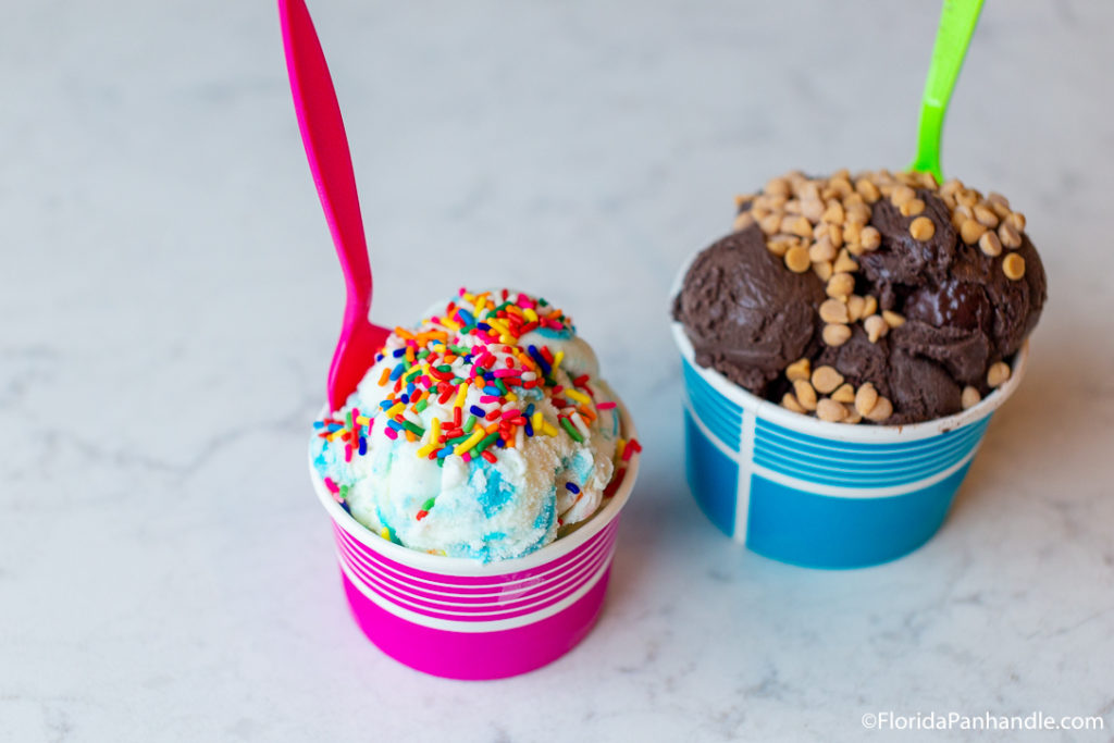 blue and vanilla swirled ice cream with rainbow sprinkles on top in a pink cup with a pink spoon next to a blue cup of chocolate ice cream with peanut butter chips on top with a lime green spoon