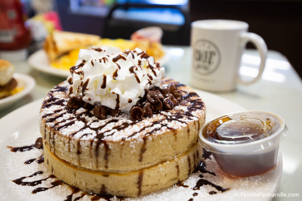 a thick stack of pancaked with powdered sugar, chocolate chips and drizzle, with whipped cream on top and a side of maple syrup