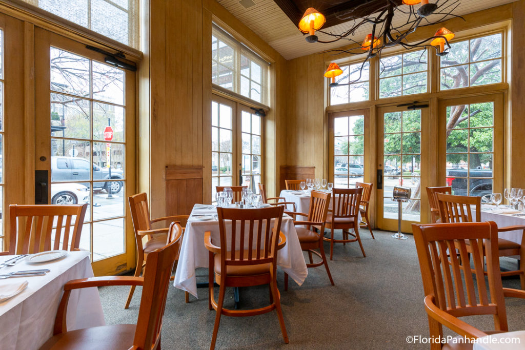 an indoor dining area with big windows and wooden chairs with white table cloths 
