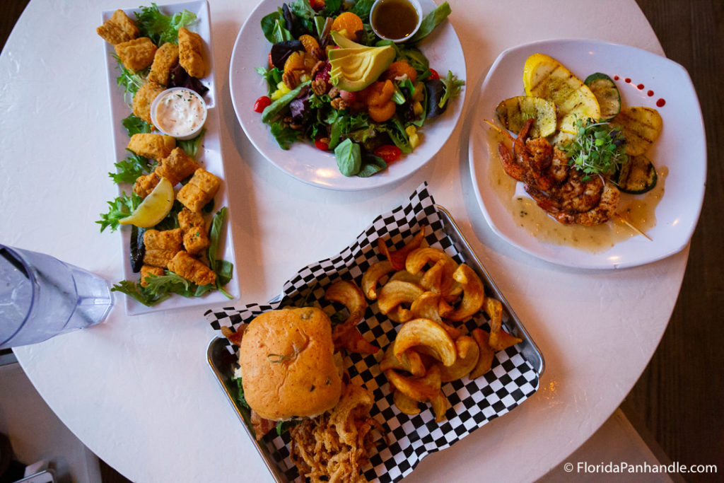 many different plates of food including, a burger with a side of fries, grilled shrimp skewers on a bed of zucchini, an avocado salad, and fish bites at Casino Beach Bar