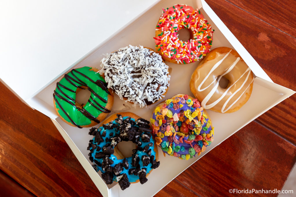 an box of 6 assorted delicious donuts - a green frosted with chocolate drizzle, a chocolate frosted with coconut flakes, a strawberry frosted with rainbow sprinkles, a blue frosted with crushed oreo chunks, a vanilla frosted with fruity pebbles cereal on top, and a maple frosted with vanilla drizzle