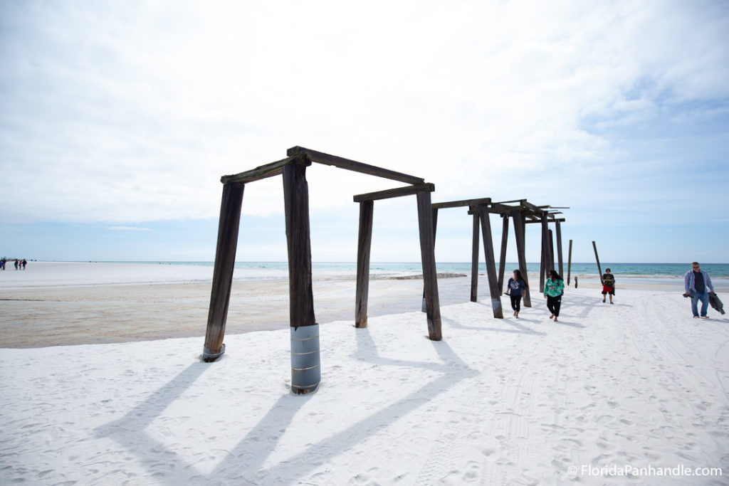 people looking around wooden structures on a white sand beach that was maybe an old pier