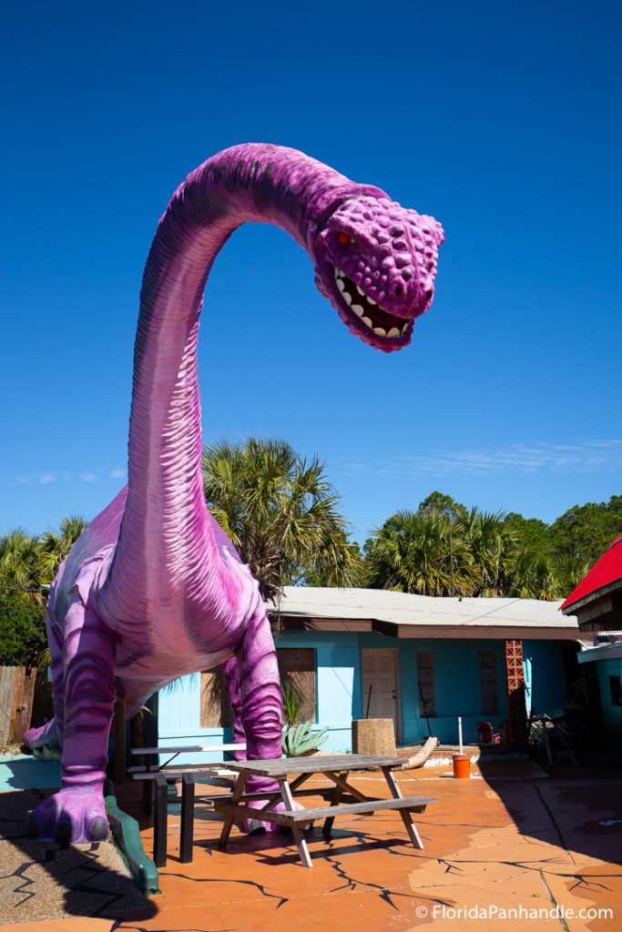 giant purple dinosaur statue stomping with long neck and red eyes on the side a small blue house and painted cracks on the floor