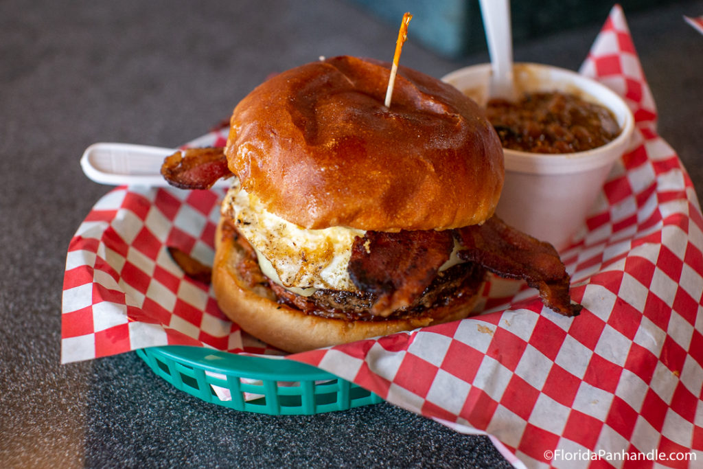 burger with fried egg and bacon on it with skewer sticking out next to styrofoam cup with chili and cheese inside - in red and white checkered basket