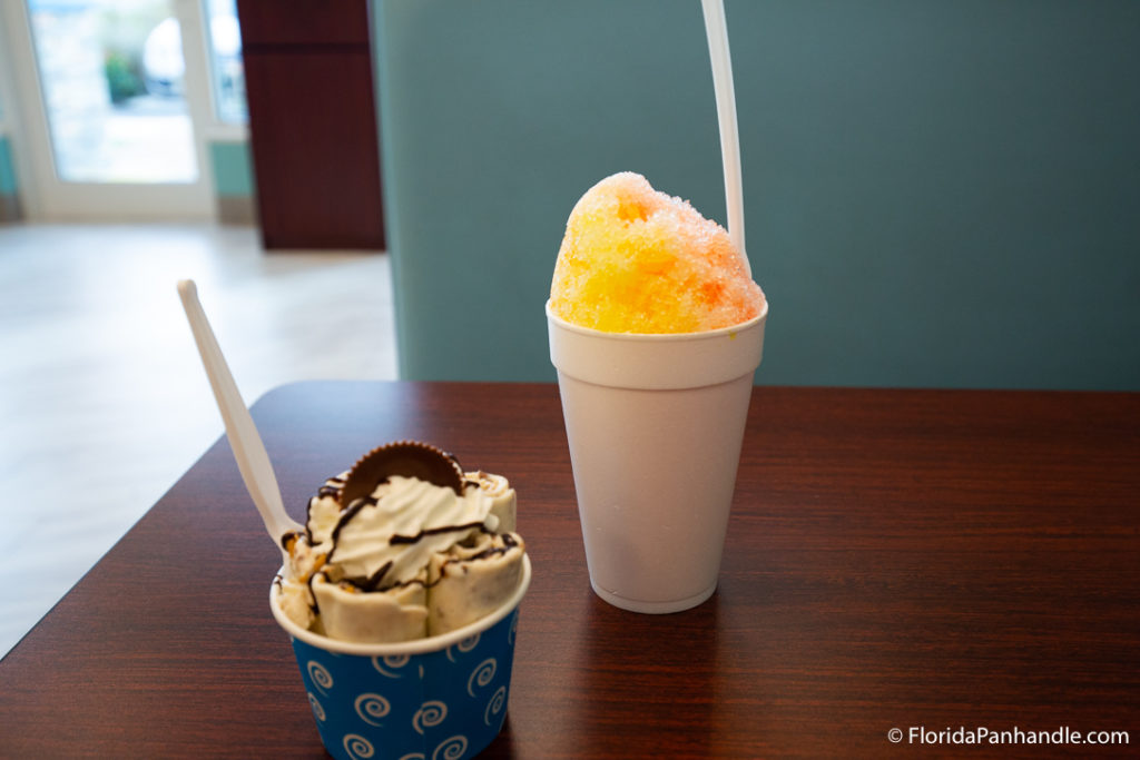a cup of rolled ice cream with whipped cream and a piece of chocolate on top next to a cup of yellow and orange shaved ice