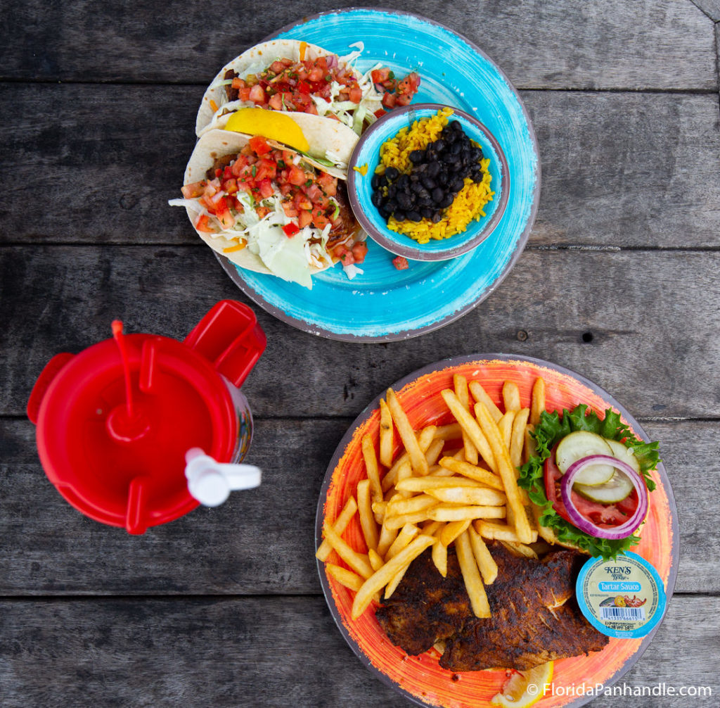 one blue plate with two tacos, rice, and black beans next to orange plate with fries and seafood with lemon on plate