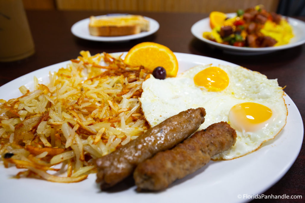 a plate of hash brown, breakfast sausage and sunny side up eggs with an orange slice on the side at The Pancakery of PCB