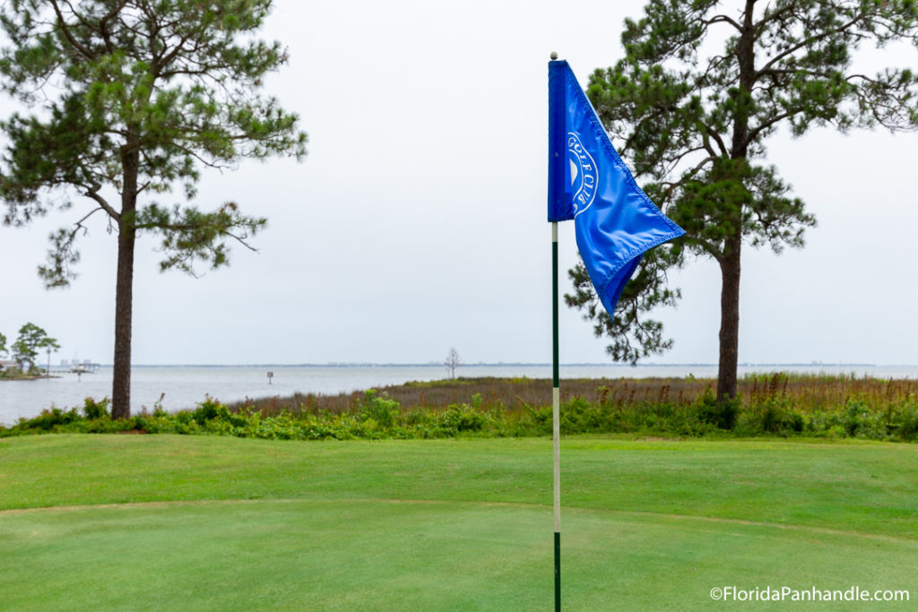 a golf pin with a blue flag surrounded by green grass and a large body of water in the background