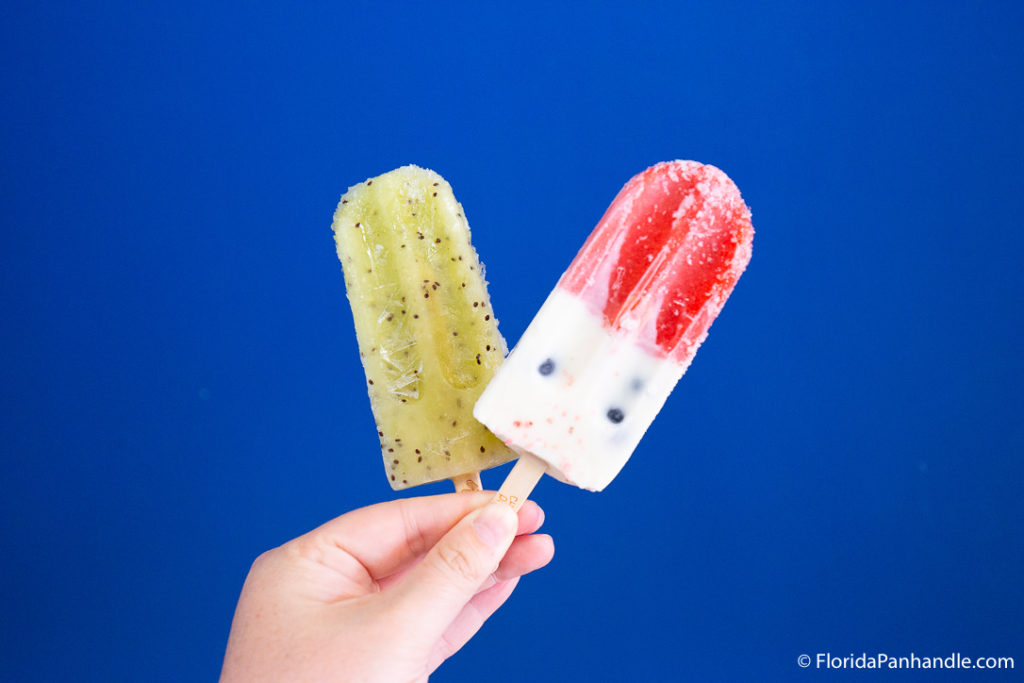 someone holding a kiwi flavored popsicle and a strawberry and vanilla flavored popsicle