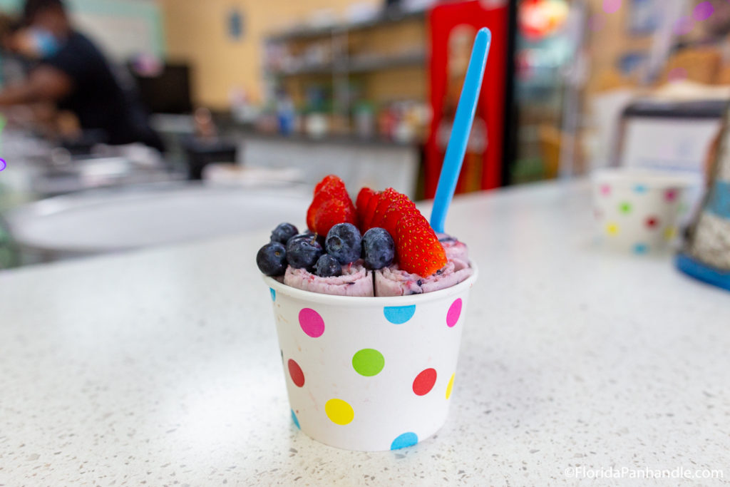 a rainbow polka dot cup filled with rolled ice cream and topped with strawberries and blueberries 