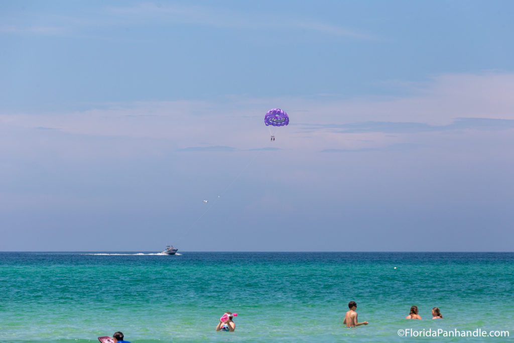people in the water and someone paragliding in the distance during the evening with bright blue and purple skies