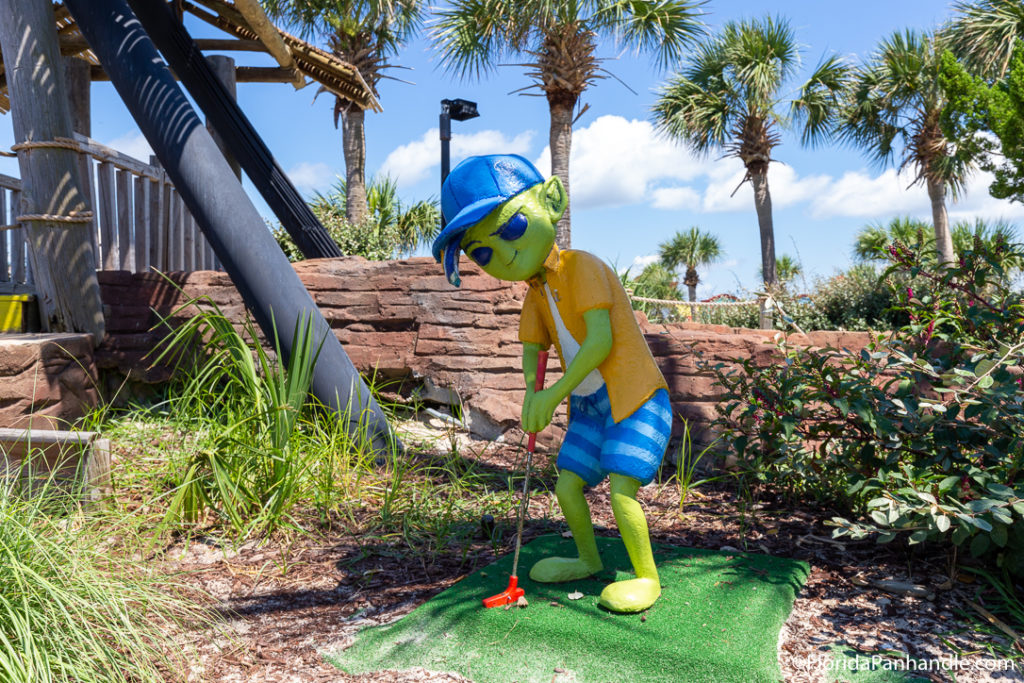 a green alien statue mini-golfing with a blue hat and beach clothes