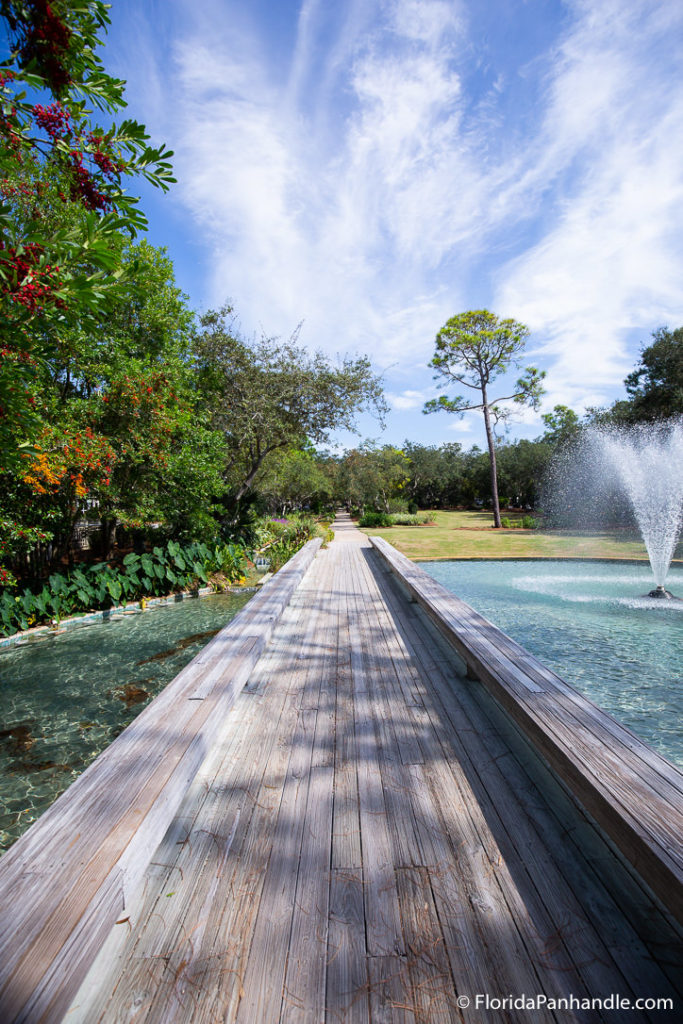 a wooden walkway over a small pond surrounded by lush greenery and some flowers