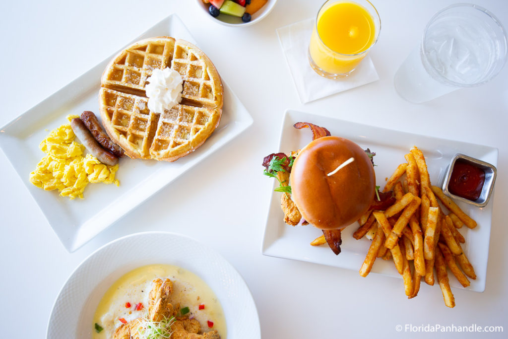 a plate of waffles with powdered sugar and whipped cream in top with a side of breakfast sausage and scrambled eggs, and a glass of orange juice