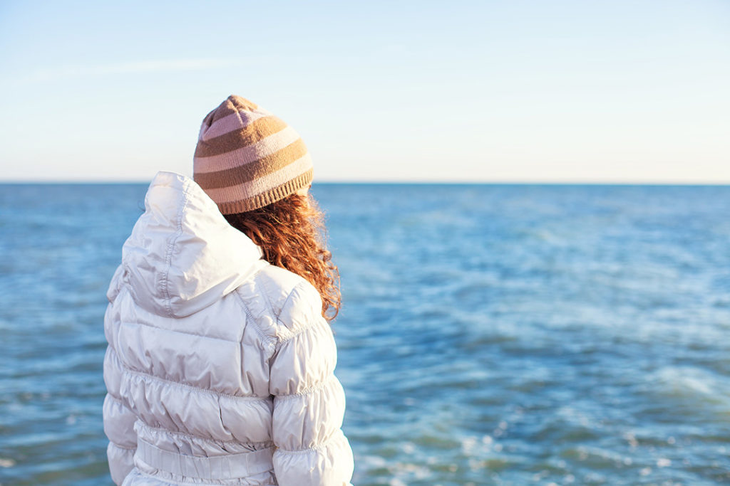 a woman wearing a white puffer jacket with a brown and baby pink stripped beanie as she faces the ocean