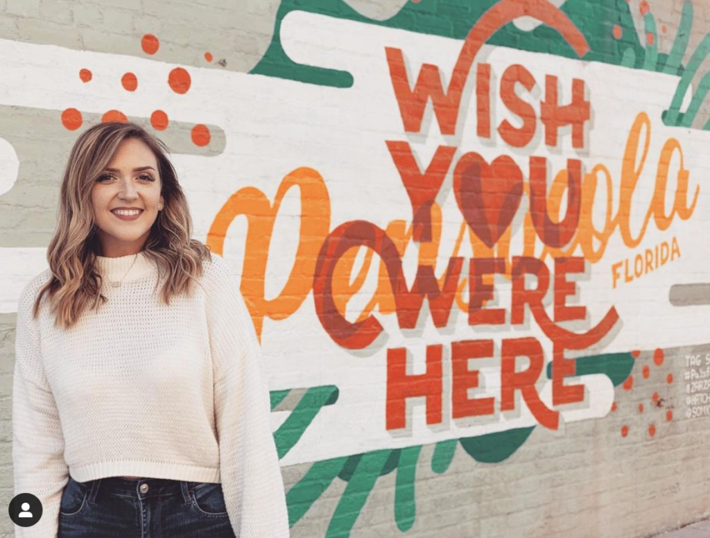 a girl standing in front of a white, green, orange, and red mural that says Wish You Were Here Pensacola Florida