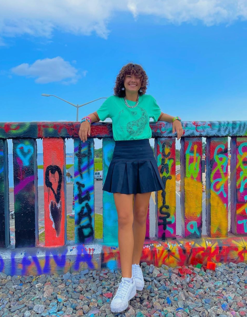 a young girl with a teal colored shirt, navy blue skirt and white shoes posing and smiling in front a railing with colorful graffiti all over it