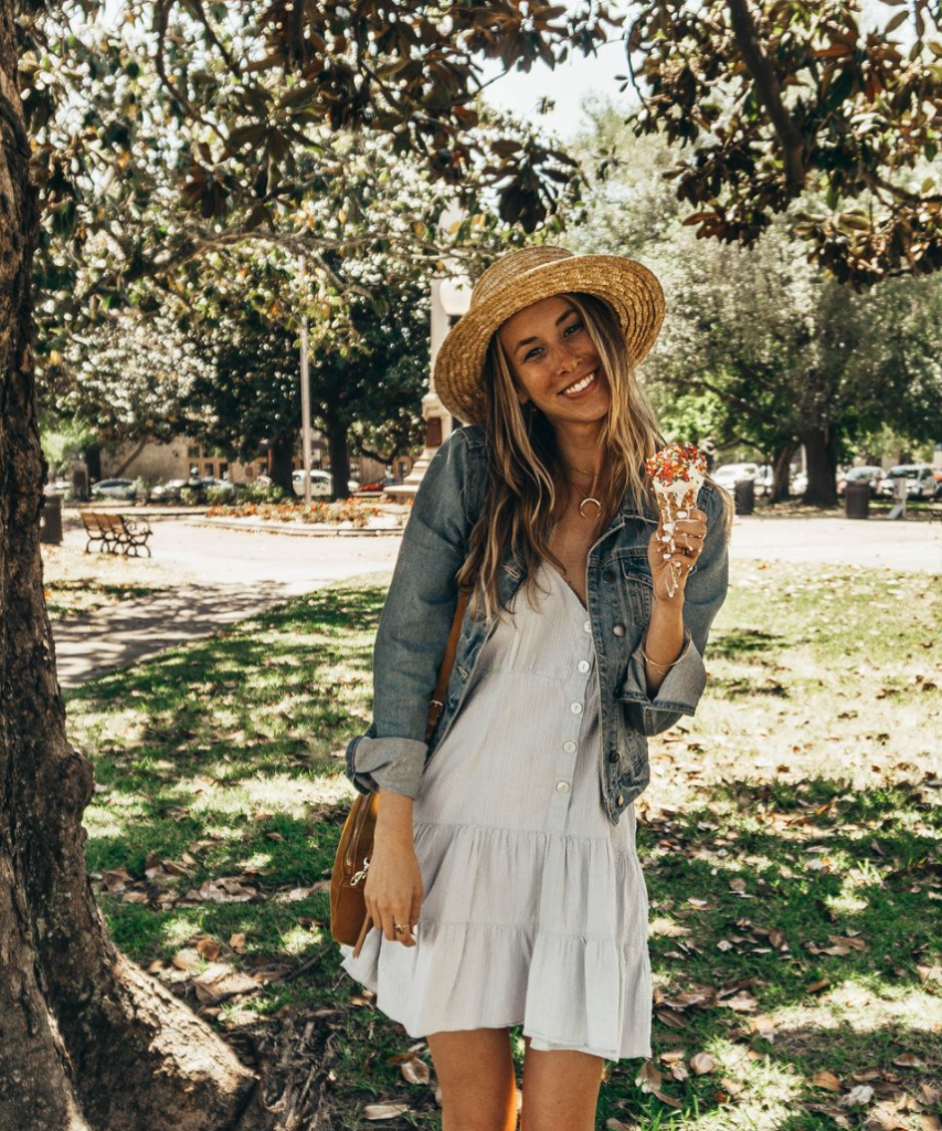 a girl with a hat and denim jacket and blue flowy dress posing and smiling under a tree while holding an ice cream cone that is melting and dripping down her fingers