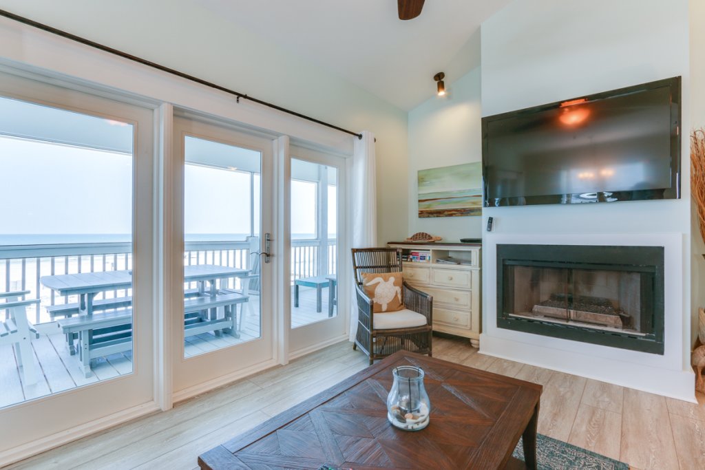 inside of a house with doors and windows facing out towards beach, wooden coffee table inside with large screen tv and fireplace